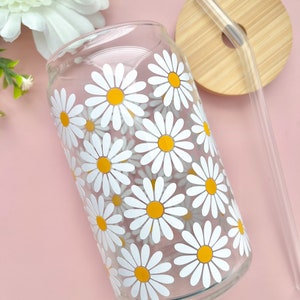 May include: A clear glass reusable can with a white daisy pattern and a yellow center. The can has a wooden lid with a clear drinking straw.
