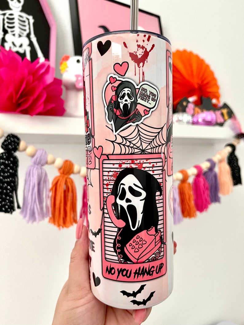May include: A pink and white stainless steel tumbler with a graphic of a ghost wearing a mask and holding a phone. The tumbler has a pink straw and the text "No you hang up" on the bottom.