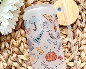 May include: A white frosted tumbler with a lid and straw. The tumbler has a variety of fall-themed illustrations, including a pumpkin, leaves, a dragonfly, a book, a coffee cup, a pizza slice, an umbrella, and ice skates. The text "YALE" is printed on the tumbler.