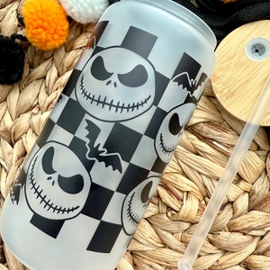 May include: A frosted glass tumbler with a black and white checkered pattern and multiple images of Jack Skellington from the Nightmare Before Christmas. The tumbler is sitting on a woven wicker surface with a clear drinking straw.