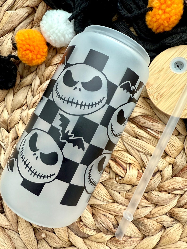 May include: A frosted glass tumbler with a black and white checkered pattern and multiple images of Jack Skellington from the Nightmare Before Christmas. The tumbler is sitting on a woven wicker surface with a clear drinking straw.