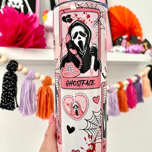 May include: A pink and white stainless steel tumbler with a Ghostface design. The tumbler features a pink phone, hearts, and a spiderweb. The text "Ghostface" is printed on the tumbler.