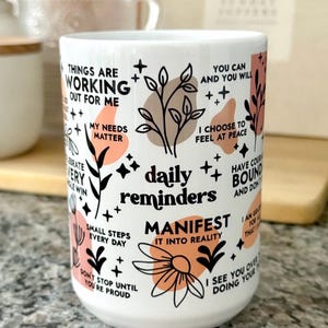 May include: White ceramic mug with the words "daily reminders" and inspirational phrases in black, peach, and brown. The mug features floral and geometric designs, including the words "Manifest it into reality."