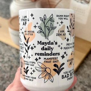 Peut inclure: Mug en céramique blanche avec le texte "Mayda's daily reminders" et diverses phrases inspirantes. Le mug présente des motifs floraux et étoilés en noir, pêche et vert. Les phrases incluent "I am strong AF" et "Manifest that shit."