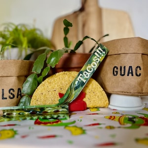 May include: A table setting for a taco party with brown paper bags labeled "SALSA", "GUAC", and a green and white ribbon that says "Tacos!!". A yellow corn tortilla is on the table with a red and green clip.