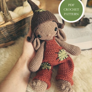 May include: Crochet pattern PDF for a brown and red gnome doll with green accents. The doll is sitting on a white fuzzy surface.