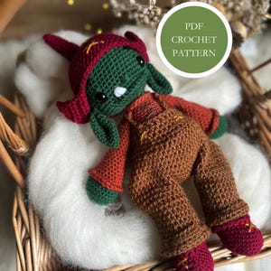 Op de afbeelding: Een handgemaakte gehaakte pop met een groen gezicht, rode muts en bruine overall. De pop heeft zwarte ogen en een kleine witte neus. De pop ligt in een rieten mand. Een groene cirkel met "PDF CROCHET PATTERN" is zichtbaar.