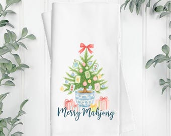 May include: A white tea towel featuring a watercolor illustration of a potted Christmas tree decorated with mahjong tiles, ornaments, and a red bow. Beneath the tree are wrapped gifts and the dark blue text that reads Merry Mahjong.