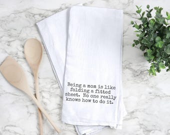 May include: White kitchen towel with the text "Being a mom is like folding a fitted sheet. No one really knows how to do it." Two wooden spoons are on the left. A green plant is on the right.