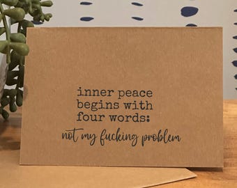 May include: A kraft paper greeting card with the text "inner peace begins with four words: not my fucking problem" in black. The card is on a wooden surface with a plant and a patterned wall in the background.