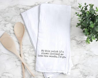 May include: White kitchen towel with the text "At this point if a clown invited me into the woods, I'd go." Two wooden spoons are on the left. A small green plant is on the right.