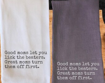 May include: Two kitchen towels, one white and one gray, are displayed. Each towel has the same text printed in black: "Good moms let you lick the beaters. Great moms turn them off first."