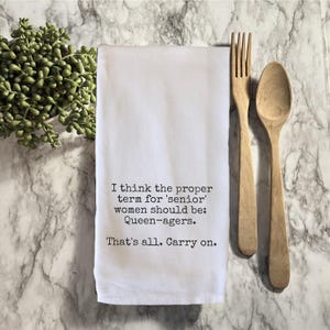May include: White kitchen towel with the text "I think the proper term for 'senior' women should be: Queen-agers. That's all. Carry on." Wooden fork and spoon are next to the towel. A small green plant is in the upper left corner.