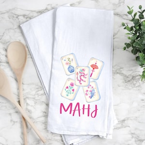May include: White kitchen towel with a watercolor design of Mahjong tiles. Each tile features a different floral or symbolic illustration. The word "MAHJ" is written in pink script below the tiles. Wooden spoons are on the left.