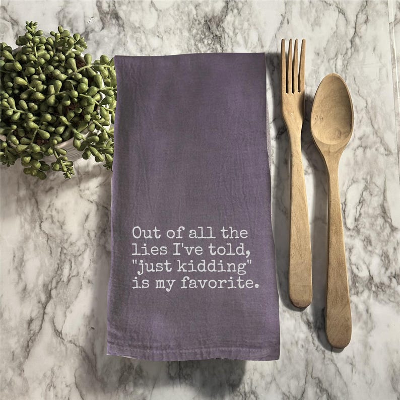 Op de afbeelding: Een paarse keukendoek met de tekst "Out of all the lies I've told, 'just kidding' is my favorite." Een houten vork en lepel liggen naast de doek. Een kleine plant in een pot staat op de achtergrond.