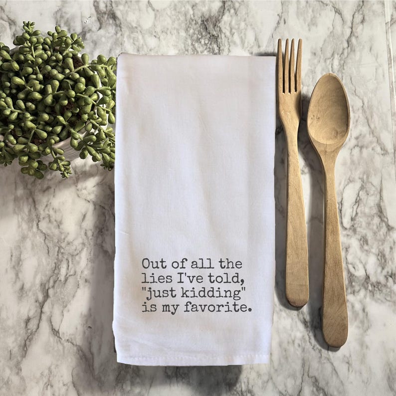 Op de afbeelding: Witte keukendoek met de tekst "Out of all the lies I've told, 'just kidding' is my favorite." vergezeld van een houten vork en lepel. Een kleine plant in een pot staat in de linkerbovenhoek. Op een marmeren oppervlak.