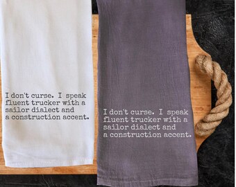 May include: Two kitchen towels, one white and one gray, are displayed on a wooden cutting board. Each towel has the text: "I don't curse. I speak fluent trucker with a sailor dialect and a construction accent."