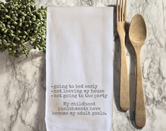 May include: White kitchen towel with black text: "going to bed early, not leaving my house, not going to the party. My childhood punishments have become my adult goals." Wooden fork and spoon are next to the towel.