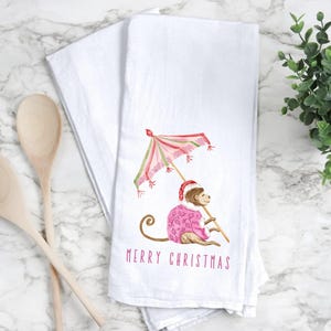 May include: White kitchen towel featuring a watercolor illustration of a monkey wearing a Santa hat and pink sweater, holding a pink and green umbrella. The text "MERRY CHRISTMAS" is printed in pink below the monkey.