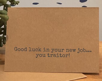 May include: A brown greeting card with the text "Good luck in your new job... you traitor!" printed in dark gray. The card is rectangular and has a simple, minimalist design, suitable for a farewell or congratulatory message.