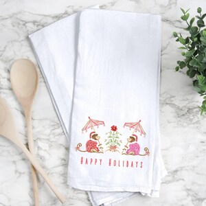 May include: White kitchen towel with a festive design featuring two monkeys under pink umbrellas, wearing holiday attire. The words "HAPPY HOLIDAYS" are printed in red below the monkeys. Wooden spoons are on the left.