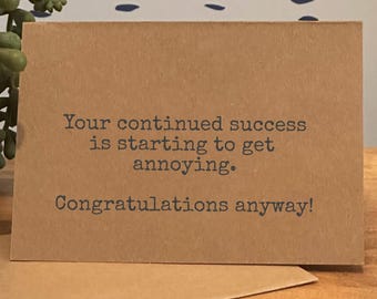 May include: A brown greeting card with the text "Your continued success is starting to get annoying. Congratulations anyway!" in dark gray. The card is on a wooden surface.