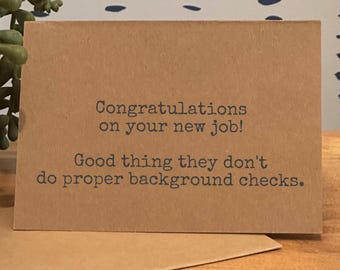 May include: A brown greeting card with the text "Congratulations on your new job! Good thing they don't do proper background checks." The card is on a wooden surface with a small succulent plant in the background.