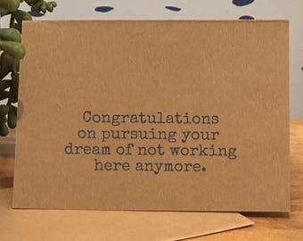 May include: A brown greeting card with the text "Congratulations on pursuing your dream of not working here anymore" in dark gray. The card is on a wooden surface.
