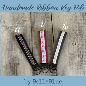 May include: Three handmade ribbon key fobs with black metal hardware. The key fobs have different ribbon designs: black with a red and white pattern, burgundy with a floral pattern, and gray with a floral pattern. The text "Handmade Ribbon Key Fob" and "by BellaBlue" are visible.