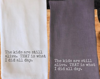 May include: Two kitchen towels, one white and one gray, with the text "The kids are still alive. THAT is what I did all day." printed in black. The towels are folded and displayed side by side.