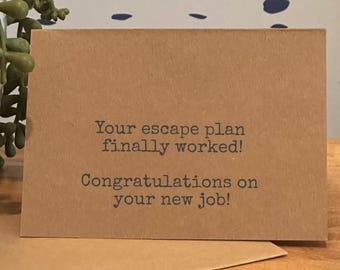 May include: A kraft paper greeting card with the text "Your escape plan finally worked! Congratulations on your new job!" in dark gray lettering. The card is on a wooden surface.