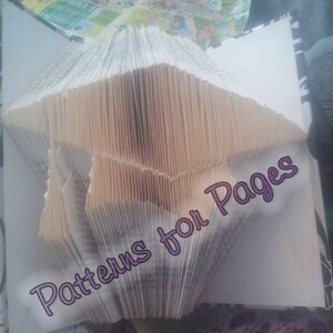 Può includere: Un libro piegato a forma di cappello di laurea. Il libro è fatto di carta bianca ed è stato piegato in una forma tridimensionale. Il testo "Patterns for Pages" è scritto in viola sul libro.