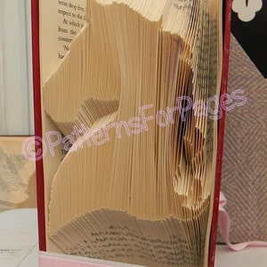 May include: A book folded into the shape of a horse. The book is bound in red and has a pink ribbon tied around it. The pages are folded into a three-dimensional shape.