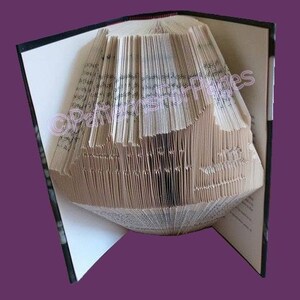 May include: A book art sculpture of a house made from the pages of a book. The pages are folded and layered to create a three-dimensional shape. The book is open and the sculpture is standing on the pages of the book.