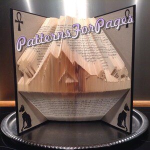 May include: A book folded into a 3D sculpture of a mountain range with two black cats on the front and back covers. The book is titled "Patterns For Pages" and features an Ankh symbol on the front cover.