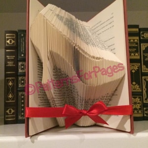 May include: A book folded into the shape of a high-heeled shoe, with a red ribbon tied around it. The book is on a shelf with other books.