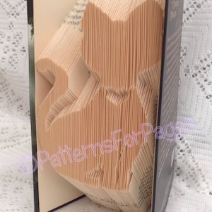 May include: A book with pages folded into the shape of a cat. The book is closed and standing upright on a white surface. The spine of the book is black and the pages are a light beige color.