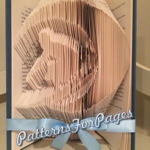 Peut inclure: Un livre dont les pages sont pliées en un design 3D d'un oiseau. Le livre est relié en bleu et a un ruban bleu clair noué autour. Le livre est intitulé "Patterns For Pages".
