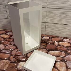 May include: A white cardboard box with a clear plastic window. The box is open and the lid is lying next to it. The box is on a background of brown stones.