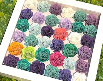 May include: A white framed shadow box filled with colorful paper flowers. The flowers are arranged in a grid pattern and are various shades of purple, green, orange, and pink. The background is a light color, and the frame is white.