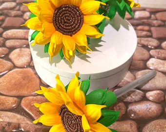 May include: Bright yellow sunflower hair accessories with brown centers and green leaves. The artificial flowers are arranged on a white surface and a stone-like surface. The sunflowers are a vibrant, cheerful design.