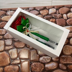 May include: A white box with a clear window, showcasing a green and red ribbon pen. The pen is decorated with a gold ribbon and a red bead.