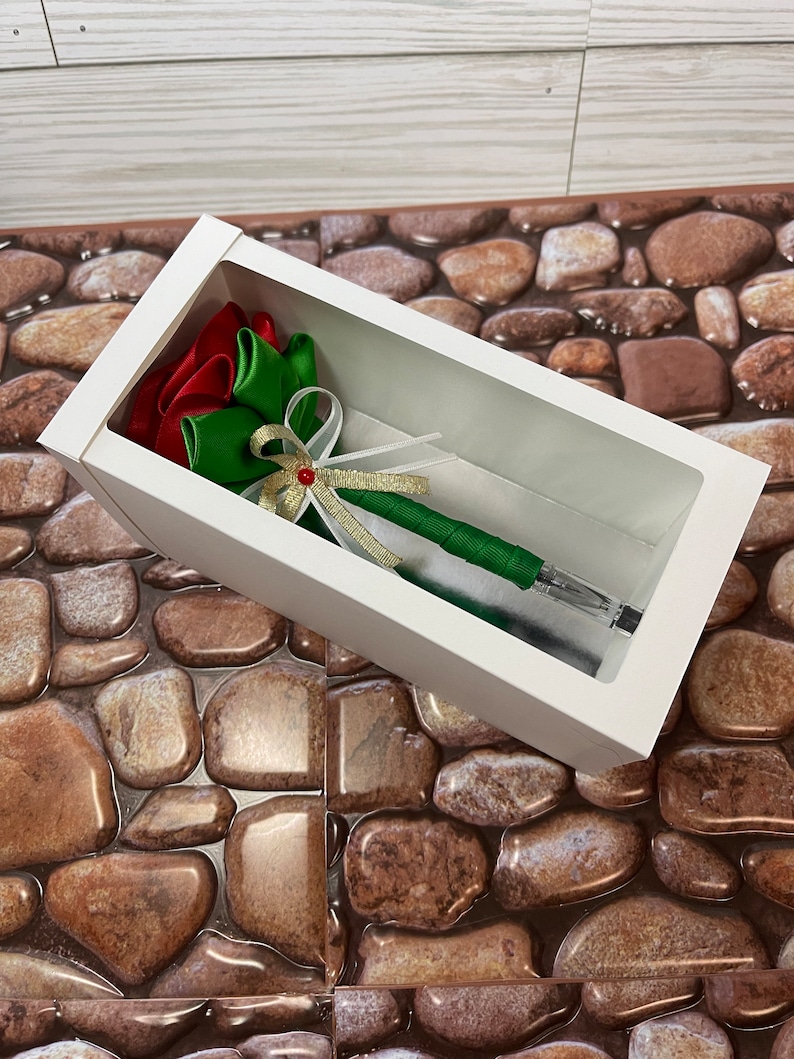 May include: A white box with a clear window, showcasing a green and red ribbon pen. The pen is decorated with a gold ribbon and a red bead.