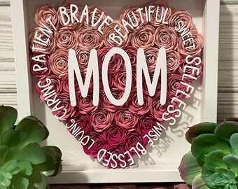 May include: A white frame with a pink and white paper flower heart that says "MOM" in the center. The heart is surrounded by words like "blessed", "wise", "loving", "caring", "patient", "sweet", "beautiful", and "brave".