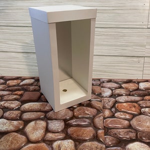 May include: A white cardboard box with an open front, revealing an empty interior. The box is sitting on a surface that looks like a stone path.