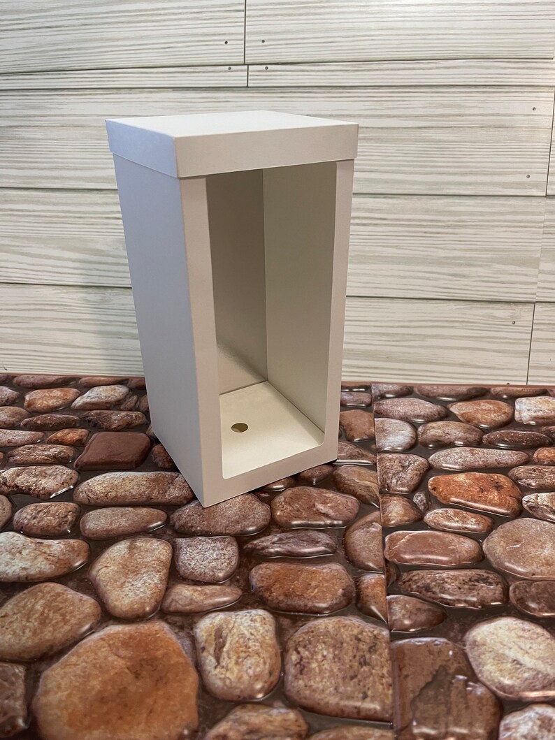 May include: A white cardboard box with an open front, revealing an empty interior. The box is sitting on a surface that looks like a stone path.