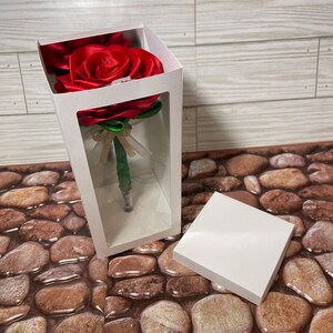May include: A red satin rose with green leaves is displayed in a clear plastic box with a white background. The rose is a pen.