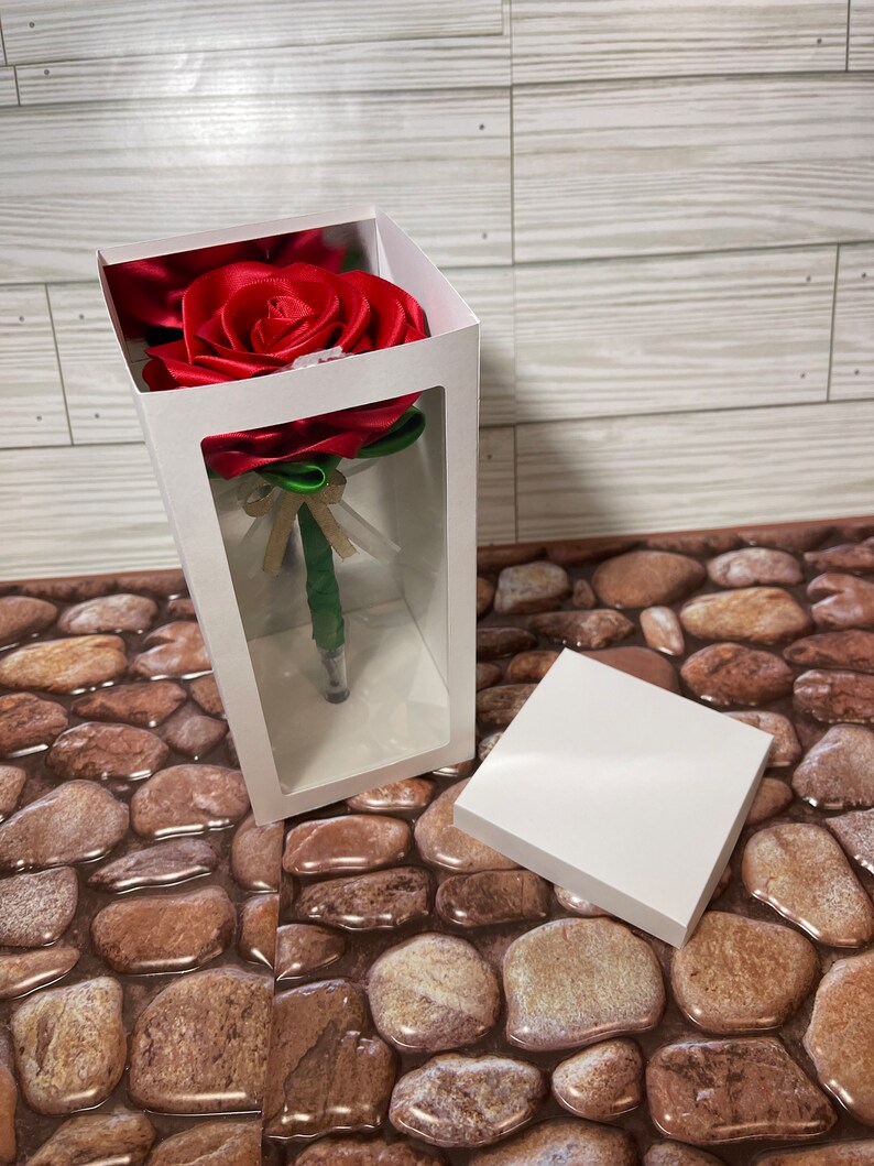 May include: A red satin rose with green leaves is displayed in a clear plastic box with a white background. The rose is a pen.