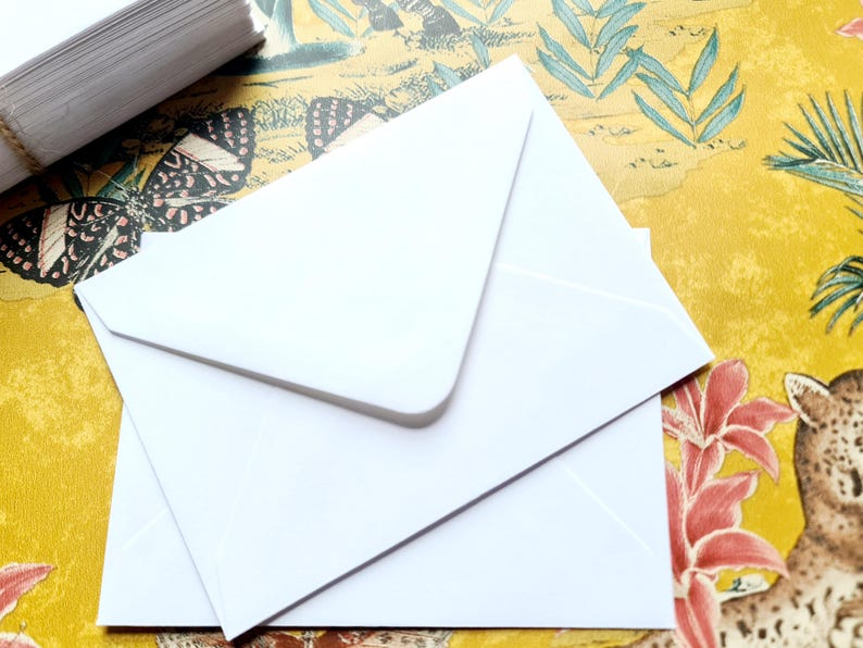May include: Two white envelopes stacked on top of each other on a yellow and green patterned surface. The envelopes are blank and have a flap on the top.