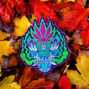 Puede incluir: Un parche bordado verde, morado y azul con un diseño psicodélico que presenta una hoja de marihuana, hongos y una cara estilizada. El parche está rodeado de hojas de otoño.